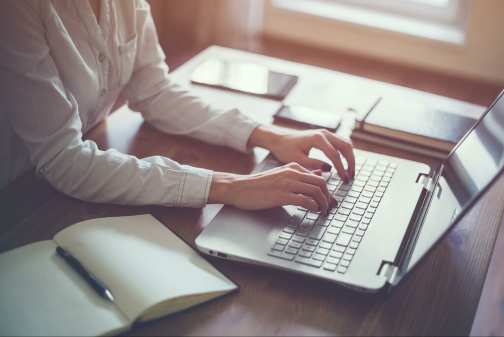 An individual in a white button down shirt types on a laptop keyboard with a stack of notebooks, a smartphone, and a tablet in the background. In the foreground, another notebook lies open with a pen resting on top of it.