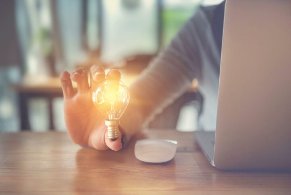 An individual reaches around a laptop to hold an illuminated light bulb between their thumb and index finger.
