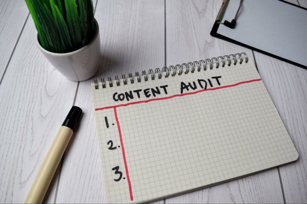 A graph paper spiral notebook lies open horizontally to show a page featuring text that reads “Content Audit” in all-caps across the top of the page and numbers 1 through 3 down the side. A black pen rests beside the notebook, as well as a potted plant and a metal clipboard.