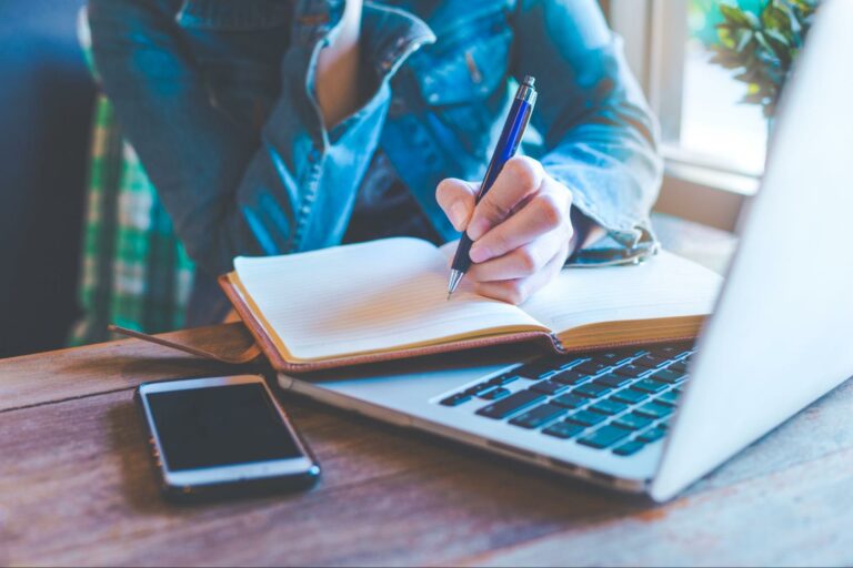 A content marketer rests a hand holding a pen on an open notebook which lies on a laptop keyboard beside a locked smartphone.