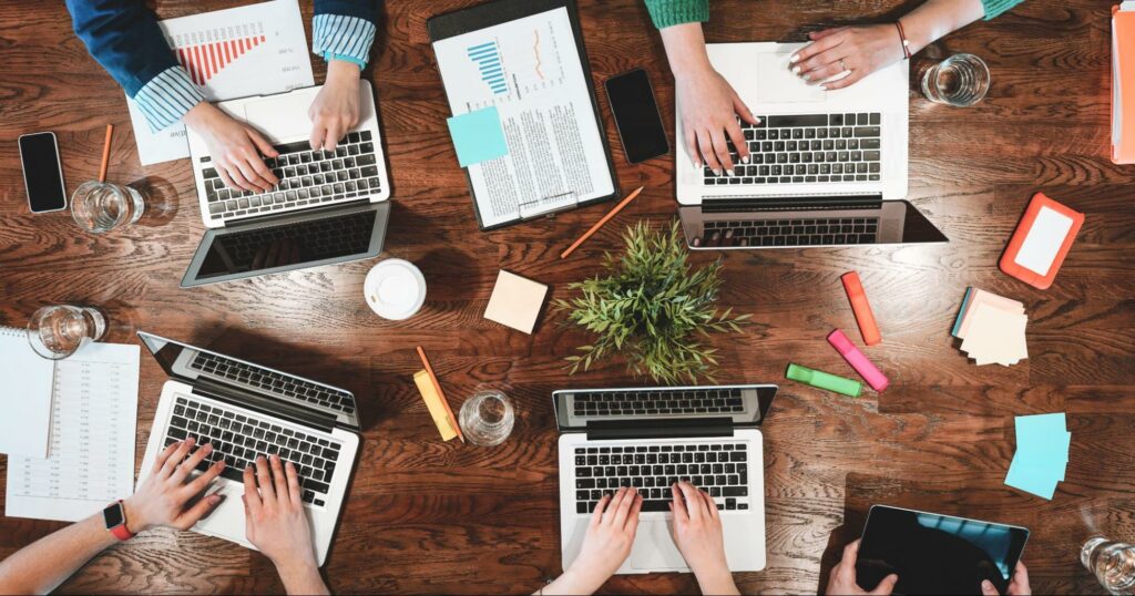 An aerial view of a large wooden table. There are four open laptops, each with a pair of hands typing on the keyboards. There are highlighters, glasses, sticky notes, graphs, and other materials spread around the table.