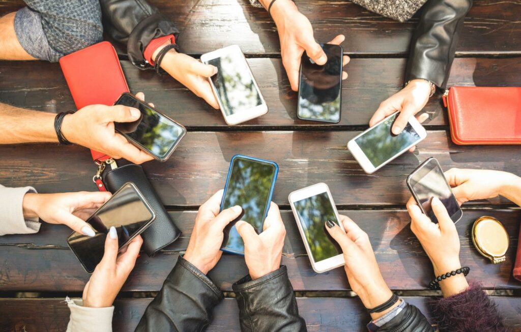 Eight individuals gather their smartphones at the center of a wooden table. While content sharing and promotion for brand awareness, lead generation, and sales purposes doesn’t look quite like this, the concept is comparable.
