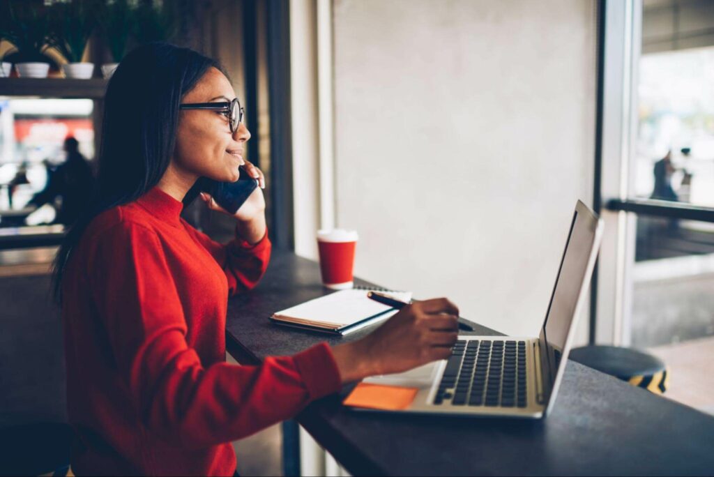 A smiling female UX copywriter in a red sweater sits in front of a laptop while speaking on her cell phone.